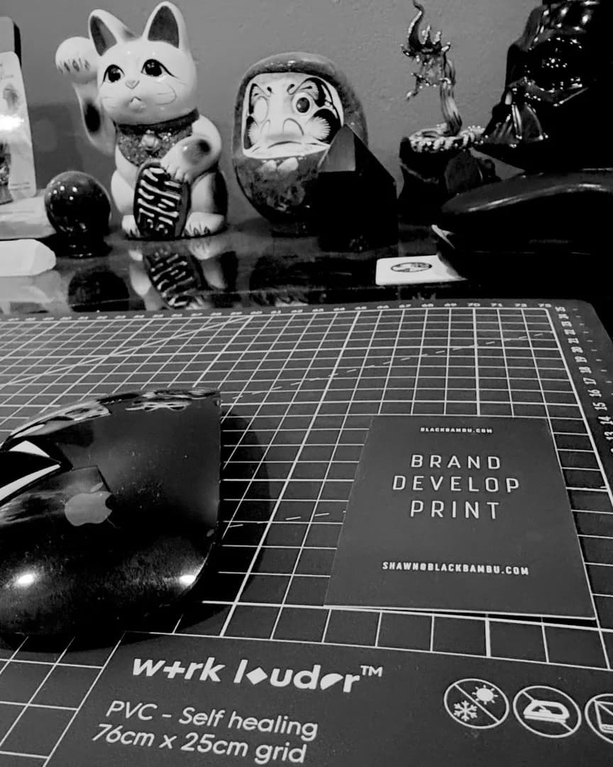Shawn’s desk with a gridded cutting mat, mouse, Black Bambu business card, lucky cat, and Daruma figure.